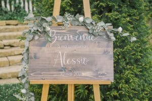 Gros plan détaillé du panneau de bienvenue en bois rustique avec l'inscription personnalisée en blanc 'Bienvenue à ma communion Alessio 10 mai 2025' et une guirlande d'eucalyptus et gypsophile a la grange aux ormes à Marly.