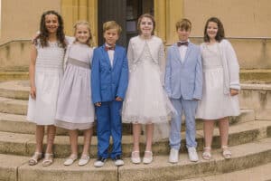 Les six enfants de Retonfey posent fièrement sur les marches en pierre devant l'entrée de l'eglise. Les quatre filles portent des robes blanches élégantes avec des accessoires de coiffure discrets, tandis que les deux garçons portent des costumes bleus (l'un bleu électrique, l'autre bleu clair) avec des nœuds papillon. Tous sont alignés et sourient face à l'objectif our leur communion a retonfey