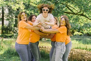 sept femmes célèbrent joyeusement un l'EVJF de Anne-Claire au jardin botanique. de montigny les metz six amies portant des t-shirts orange et des lunettes de soleil en forme de cœur soulèvent dans les airs la future mariée, vêtue d'un t-shirt blanc, qui saute avec enthousiasme. Elles rient toutes devant un arrière-plan verdoyant d'arbres et de fleurs
