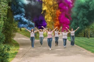 Cette photo en mouvement, prise en perspective sur un chemin pavé du parc thermal de Mondorf-les-Bains, capture les six participantes de l'EVJF de Perrine sortant d'une haie d'honneur de fumigènes colorés. La mariée Perrine, en t-shirt rose au centre, et ses amies, en t-shirts blancs "Team de la mariée", ont les bras levés en signe de joie et de triomphe