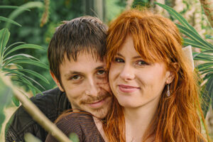 Gros plan intime de Léna et Paul, de la love session, leurs visages rapprochés, souriant à l'objectif, encadrés de manière floue par des feuilles de plantes grasses au premier plan