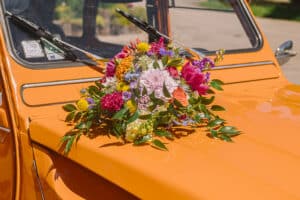 Gros plan d'un bouquet de fleurs champêtres aux couleurs vives (jaune, rouge, violet) posé sur le capot d'une 2CV orange vintage pour le mariage de Julie et Christophe