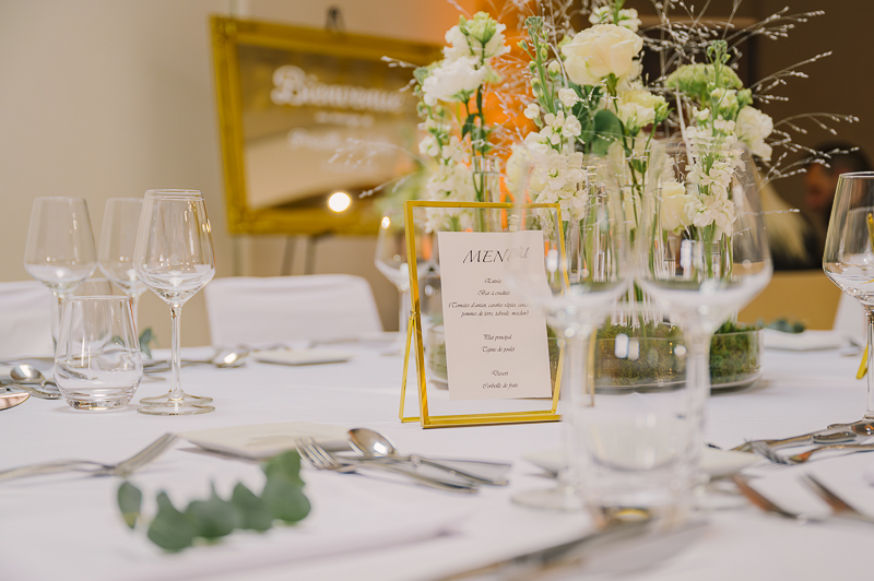 Gros plan sur un menu de mariage élégamment présenté dans un cadre doré, au centre d'une table dressée par 8Mdeco pour le salon de Luttange. Le menu détaille les plats de David Ciré Traiteur (Entrée : Bar à crudités... Plat : Tajine de poulet... Dessert : Corbeille de fruits). Des verres à pied en cristal et des arrangements floraux délicats de fleurs blanches et feuillage d'Et l'eau dit encadrent le menu, posé sur une nappe blanche avec des couverts argentés