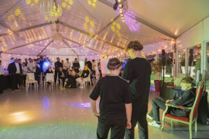 Vue en perspective depuis l'arrière de la piste de danse. De dos, deux jeunes freres (les animateurs) regardent vers le fond du chapiteau où tous les invités sont réunis et écoutent. Lorenzo est visible à droite, toujours assis dans son fauteuil rouge. Le chapiteau est décoré de lustres et de lumières de fête.