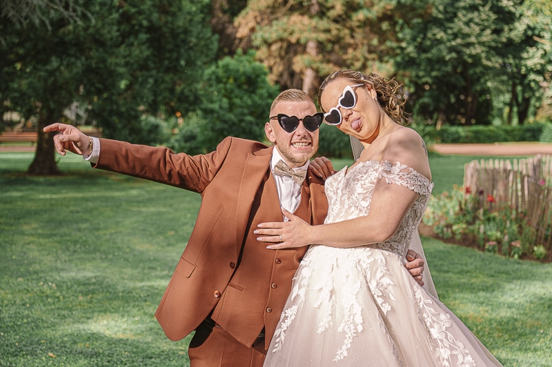 Photo de mariage drôle et décalée du couple portant des lunettes de soleil blanches et noires en forme de cœur, Charlène faisant une grimace amusée en tirant la langue.