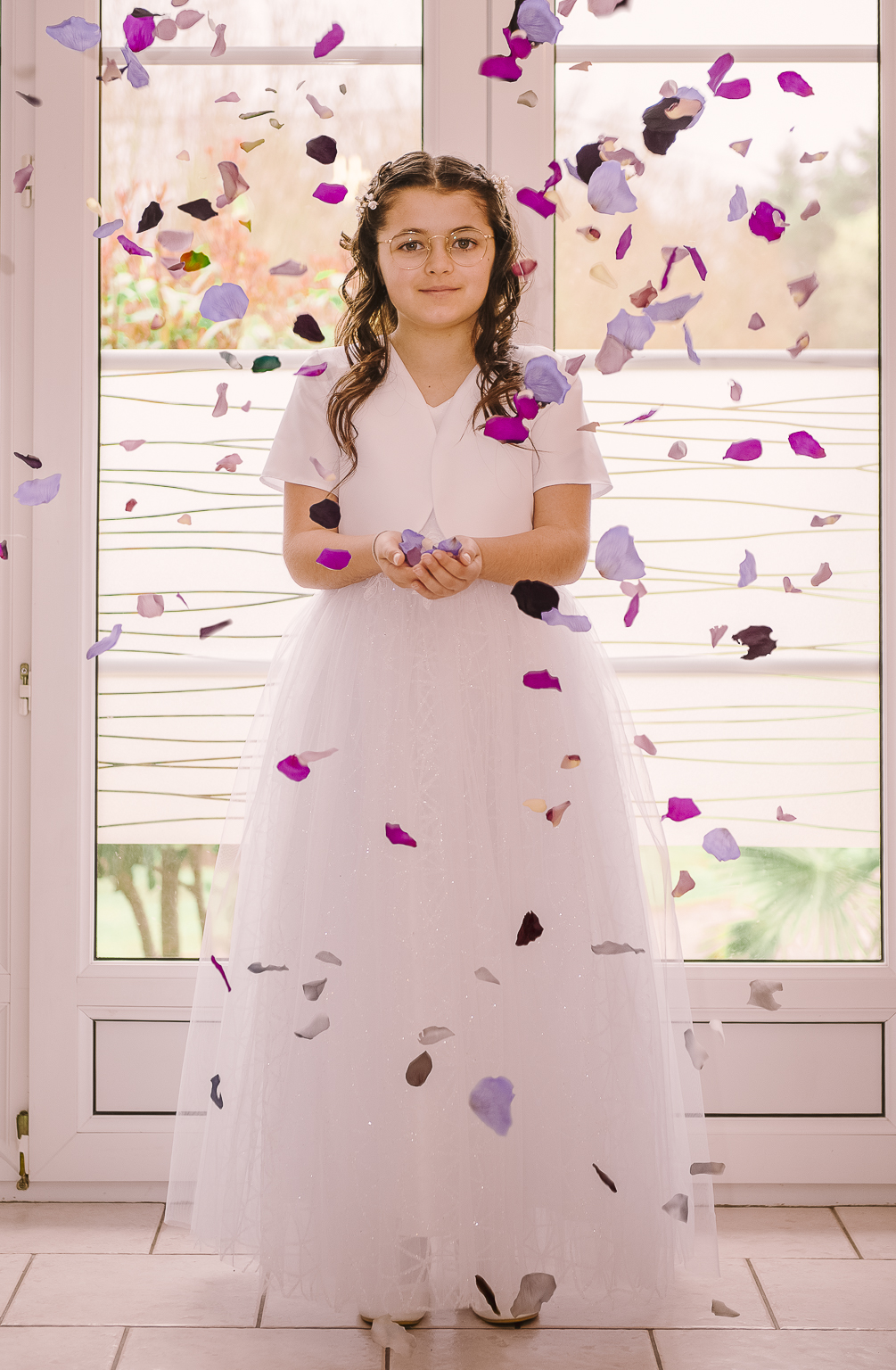 Portrait en pied d'une jeune fille en robe de communion blanche et veste assortie, avec lunettes, debout devant une porte-fenêtre sous une pluie de pétales de fleurs violettes, bleues et noires.