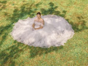 Prise de vue aérienne de la mariée assise sur l'herbe déployant la traîne en tulle de sa robe de mariée.