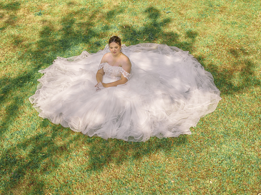Prise de vue aérienne de la mariée assise sur l'herbe déployant la traîne en tulle de sa robe de mariée.